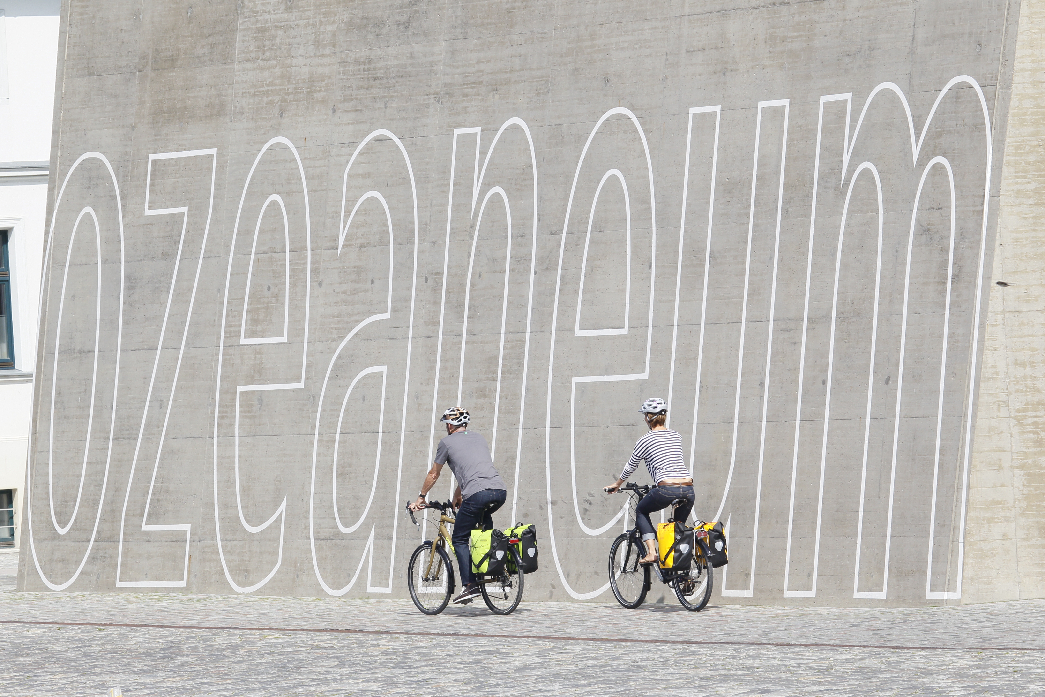 Zwei Radfahrer mit dem Rücken zur Kamera fahren mit ihren Fahrrädern an einer Wand vorbei. Auf der Wand steht in großer weißer Schrift &#34;OZEANEUM&#34;